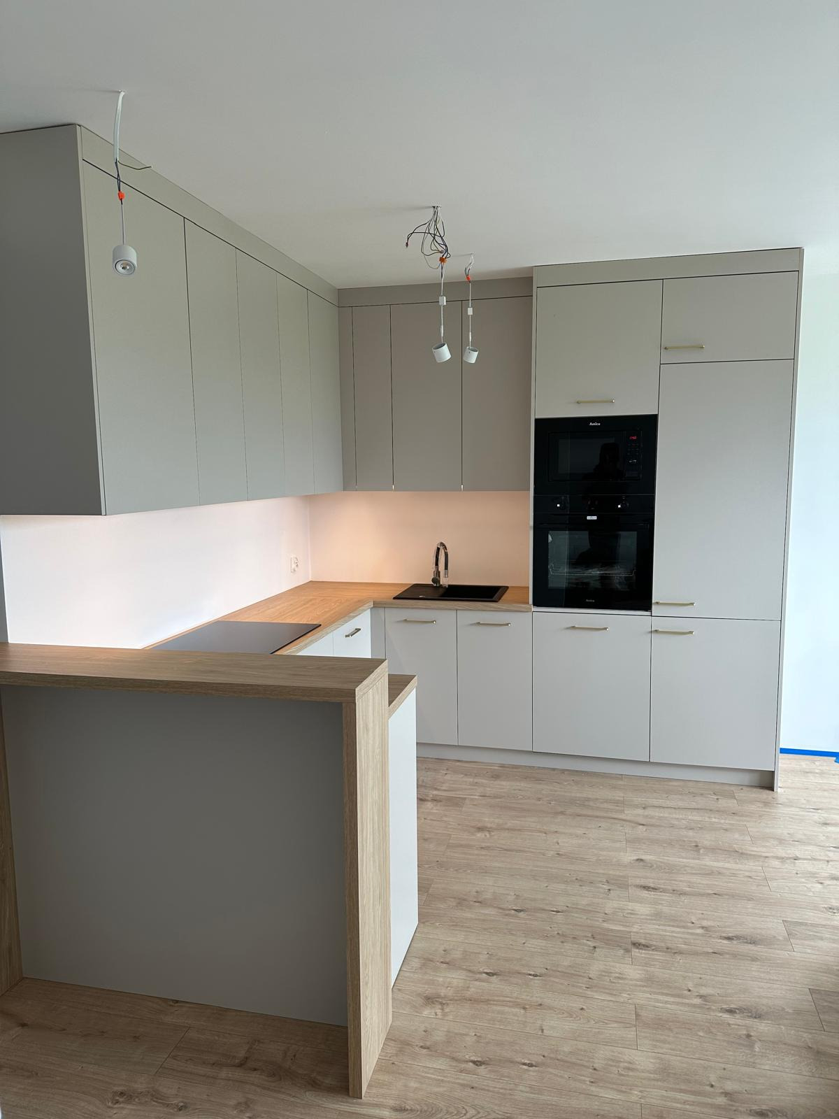 Jasna kuchnia z drewnianym blatem i barkiem, oświetlenie podszafkowe i wiszące lampy bez kloszy na kablach.