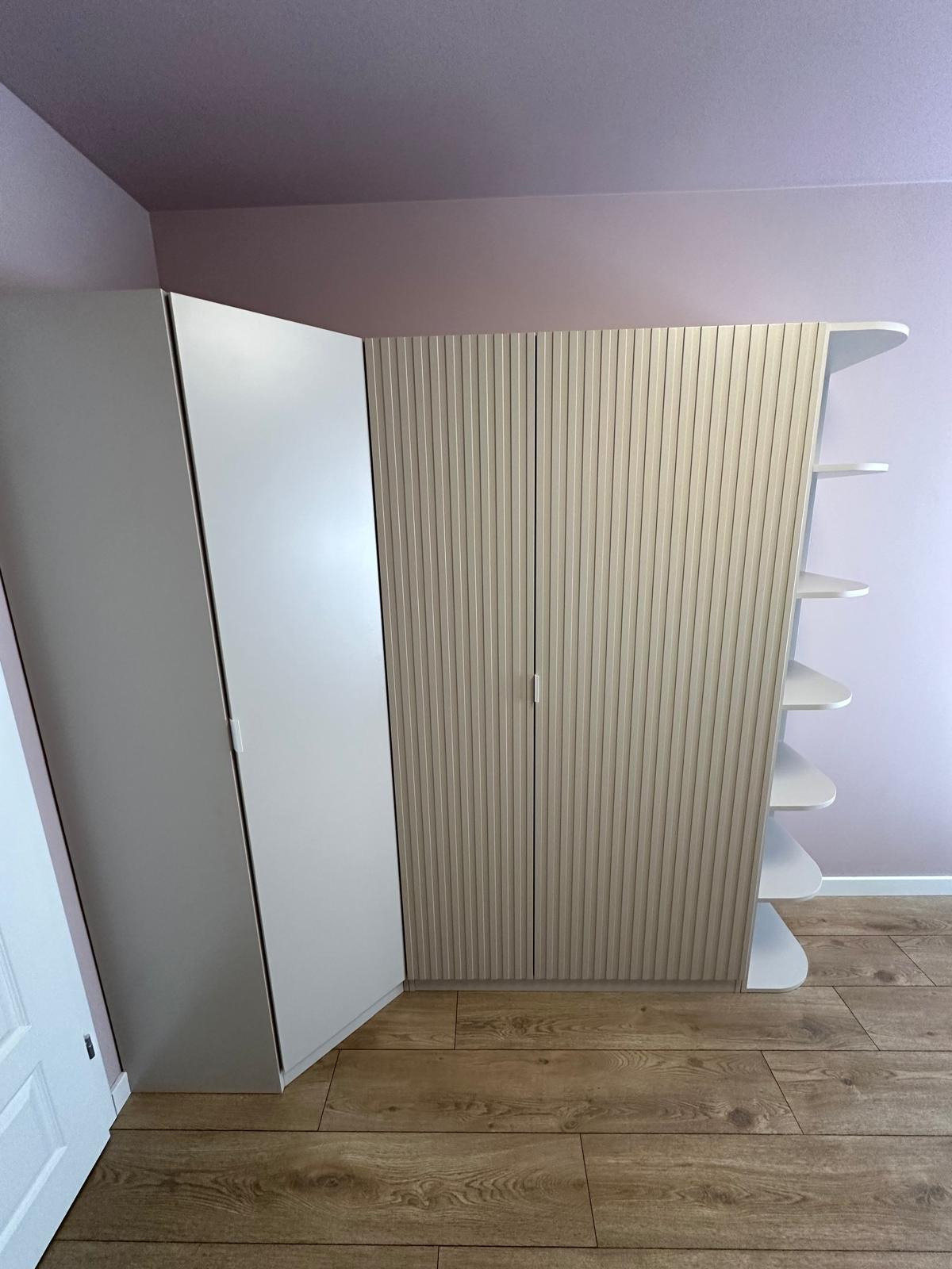 Szafa wnękowa z białymi frontami i ryflowanymi drzwiami w odcieniach beżu, z otwartymi półkami na boku, na drewnianej podłodze, ściana w kolorze lila.