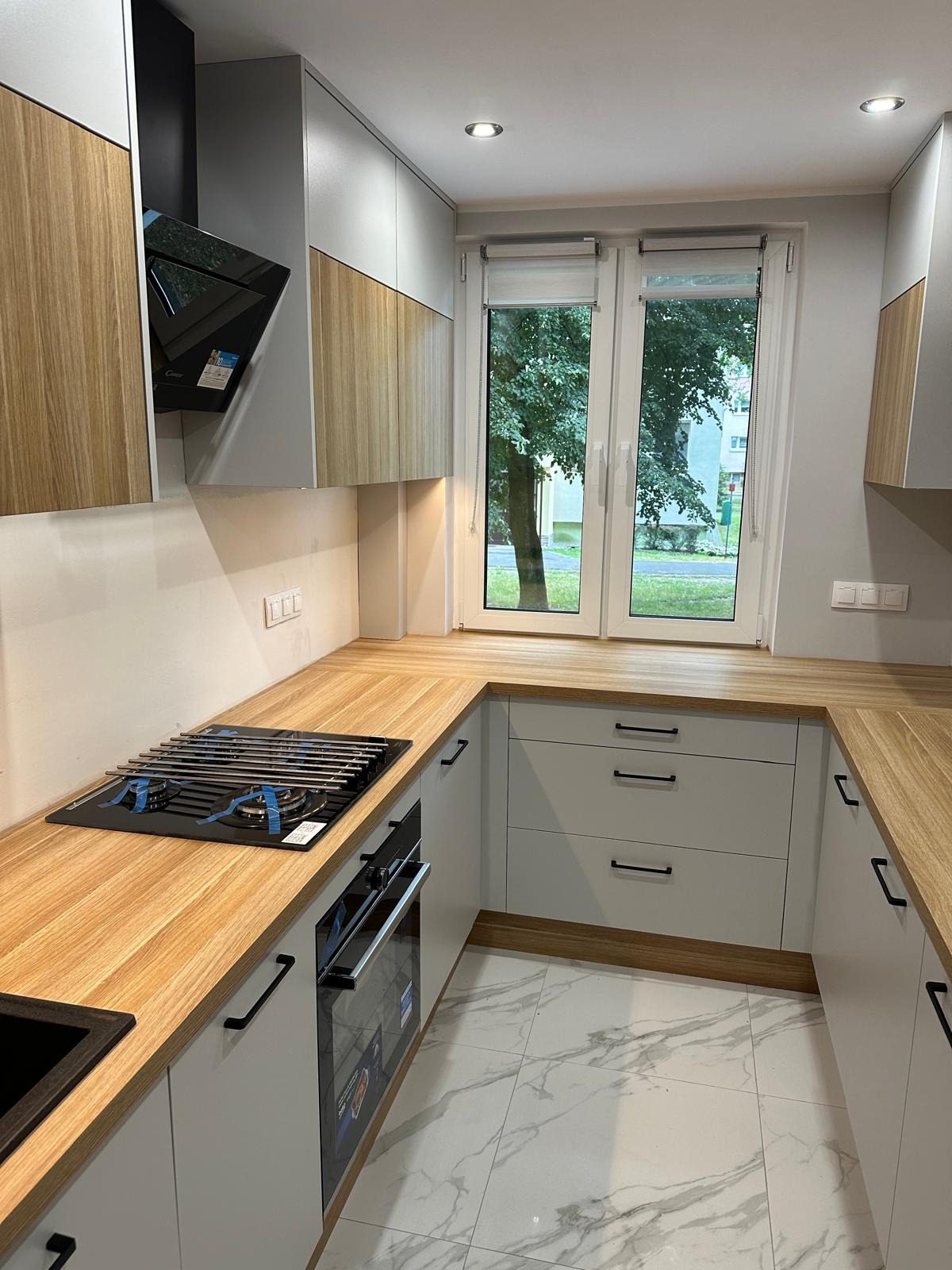 Jasna kuchnia z drewnianym blatem, białymi szafkami z czarnymi uchwytami, marmurową podłogą i widokiem na zieleń za oknem. Nowa płyta gazowa zabezpieczona folią.