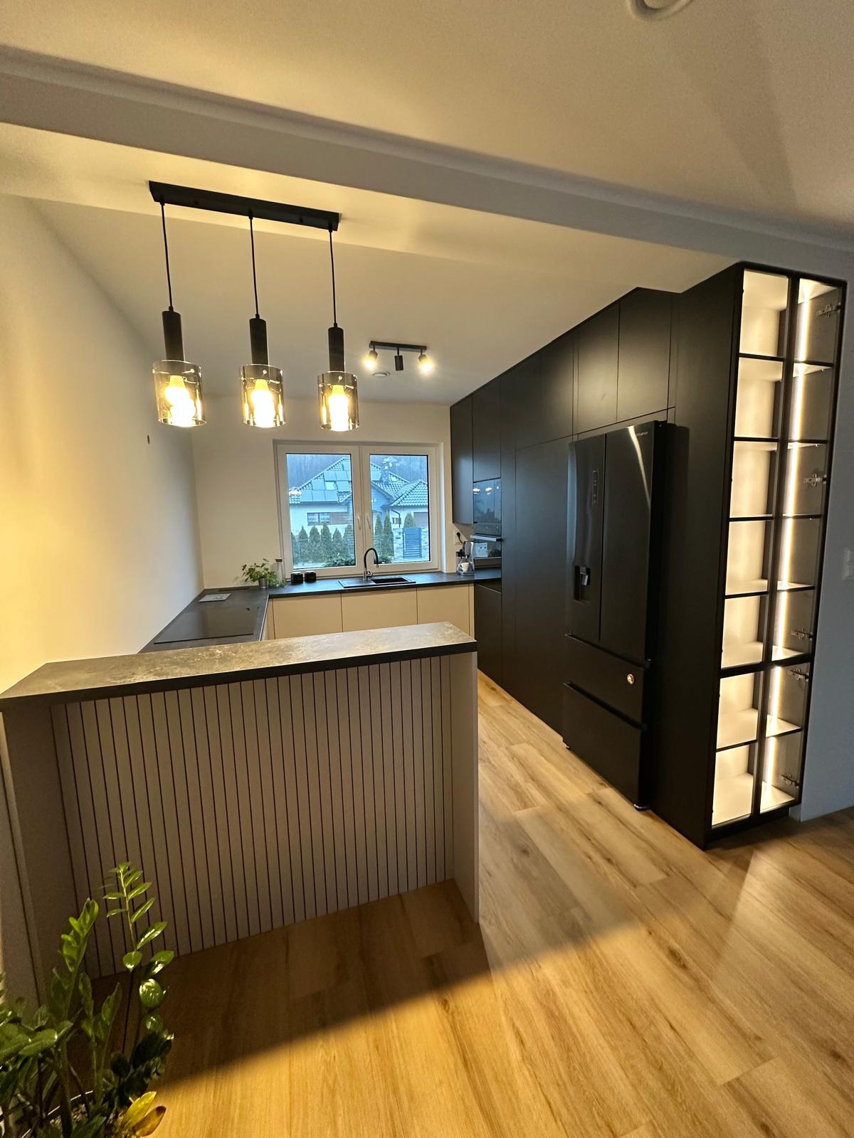 Nowoczesna kuchnia z czarnymi szafkami, wyspą z pionowymi panelami i oświetloną witryną, uzupełniona drewnopodobną podłogą i trzema wiszącymi lampami.