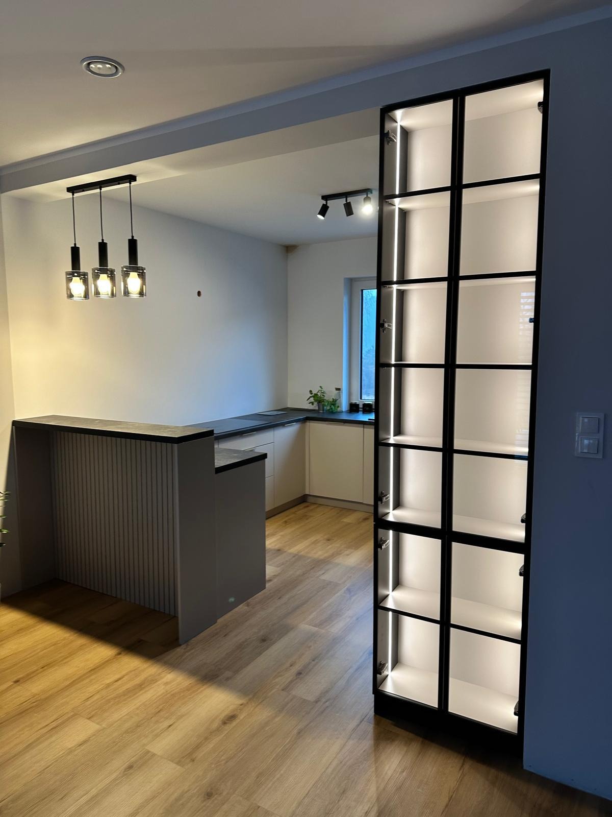 Nowoczesna kuchnia z barkiem i wysoką szafą wnękową z podświetleniem LED, widok na drewnianą podłogę