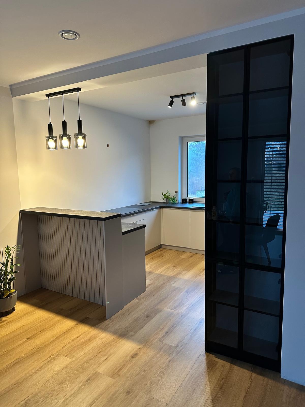 Nowoczesna kuchnia z wyspą i czarną, przeszkloną ścianką działową, oświetlona trzema wiszącymi lampami z kwadratowymi kloszami, z widocznym fragmentem białych szafek i drewnianą podłogą.