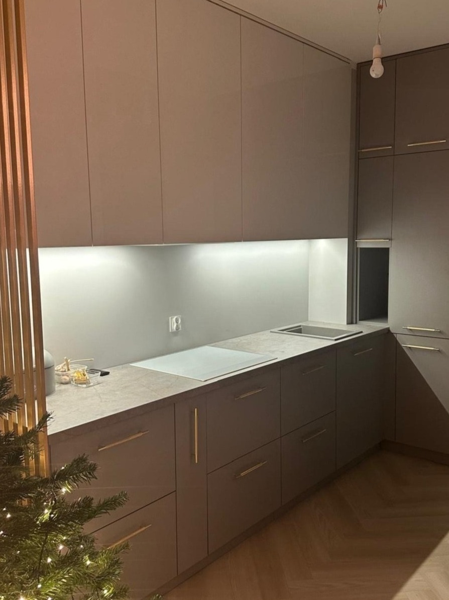 Szara, minimalistyczna kuchnia z podświetleniem LED pod szafkami, blatem imitującym kamień i drewnianą podłogą w jodełkę, częściowo zasłonięta przez świąteczne drzewko.