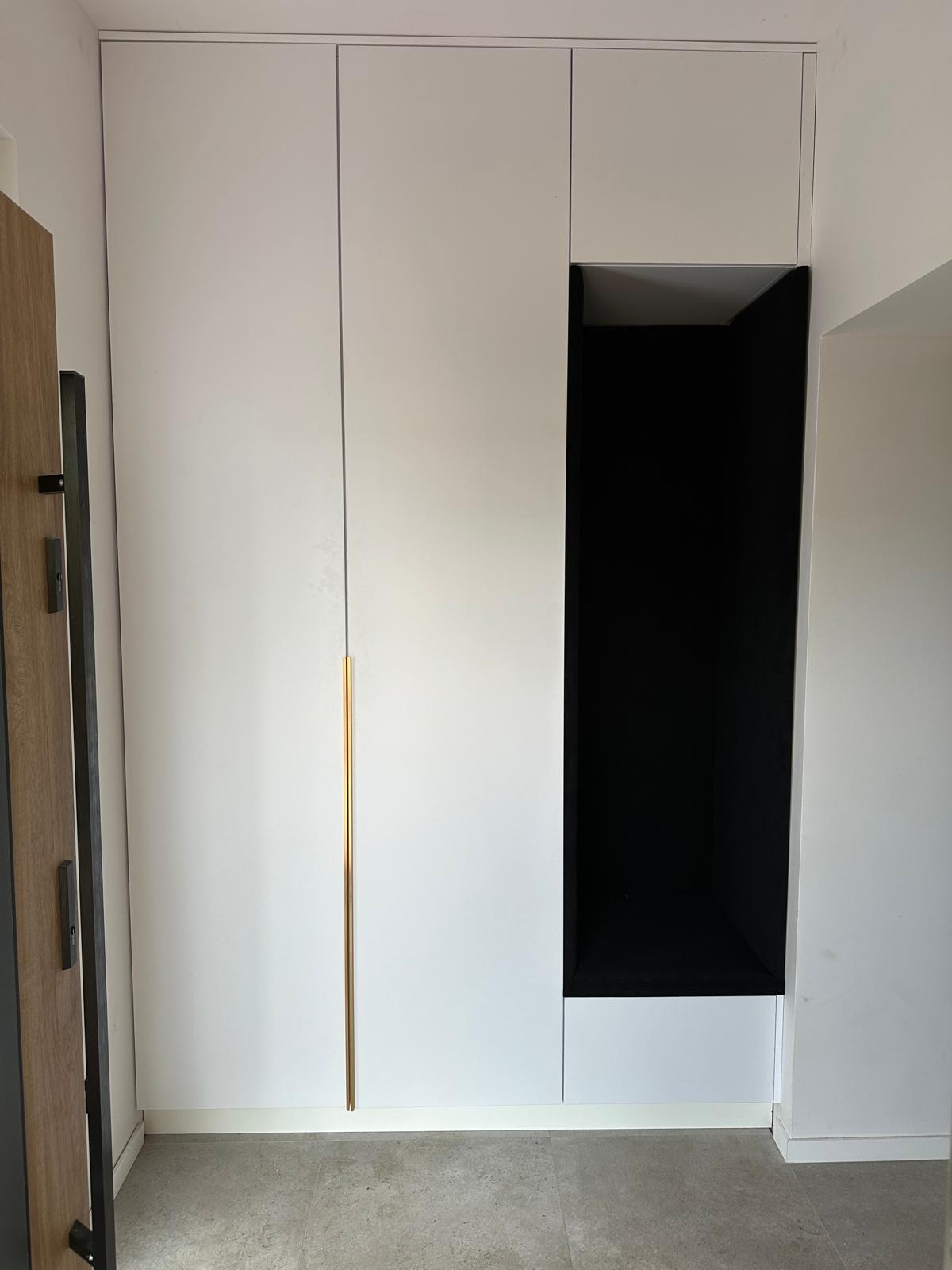Biała szafa wnękowa z długim złotym uchwytem i otwartą czarną półką, widoczna część drzwi po lewej stronie.