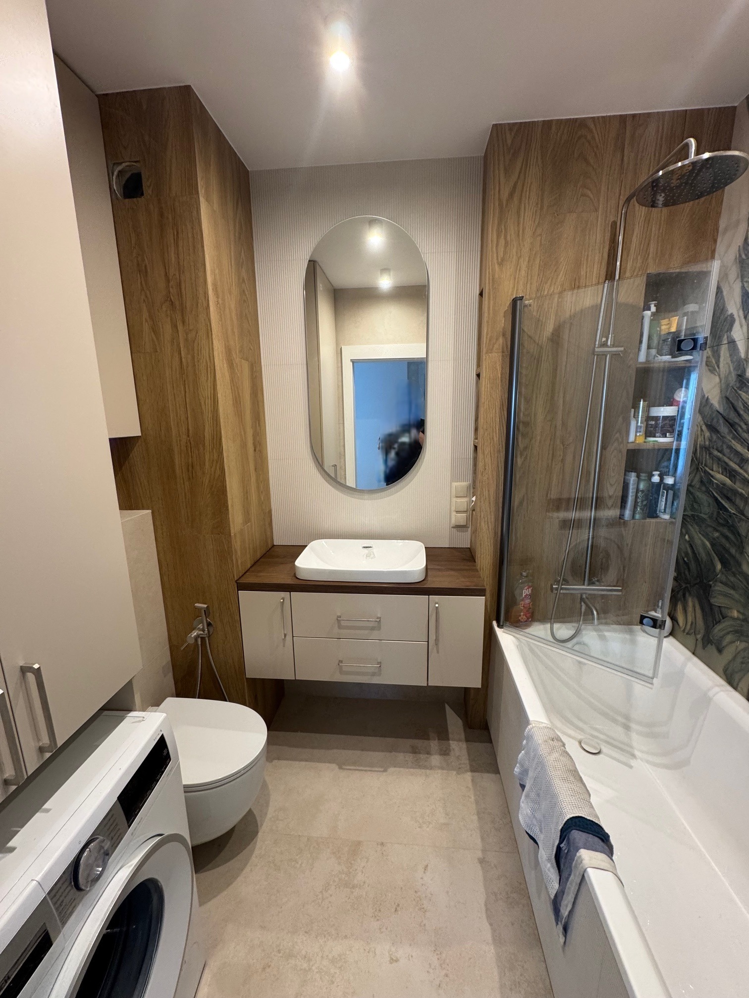 Nowoczesna łazienka z drewnianymi akcentami: umywalka z szafką, wanna z parawanem, toaleta i pralka. Owalne lustro odbija wnętrze. Minimalistyczny design.