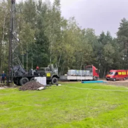 Wiertnica na podwoziu ciężarówki, dwóch pracowników przy pracy, obok ciężarówka z pojemnikami na wodę i czerwony bus z logo firmy na tle drzew i zielonej trawy.