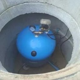 Waterwell - Daniel Janusz - Niebieski zbiornik hydroforowy z manometrem i elementami instalacji w betonowym kręgu studni. Widoczne przewody i złączki.