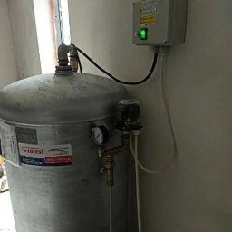 Waterwell - Daniel Janusz - Szary zbiornik ciśnieniowy z manometrem i podłączeniami hydraulicznymi, zasilany z puszki elektrycznej z zieloną diodą, widoczny w jasnym pomieszczeniu.
