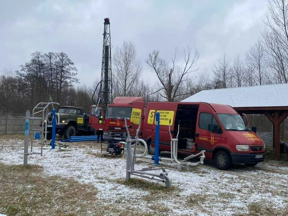 Wiercenie studni głębinowej: czerwony samochód ciężarowy z wieżą wiertniczą na zasnieżonym terenie, obok zaparkowany czerwony bus z otwartymi drzwiami i sprzętem do ćwiczeń na zewnątrz