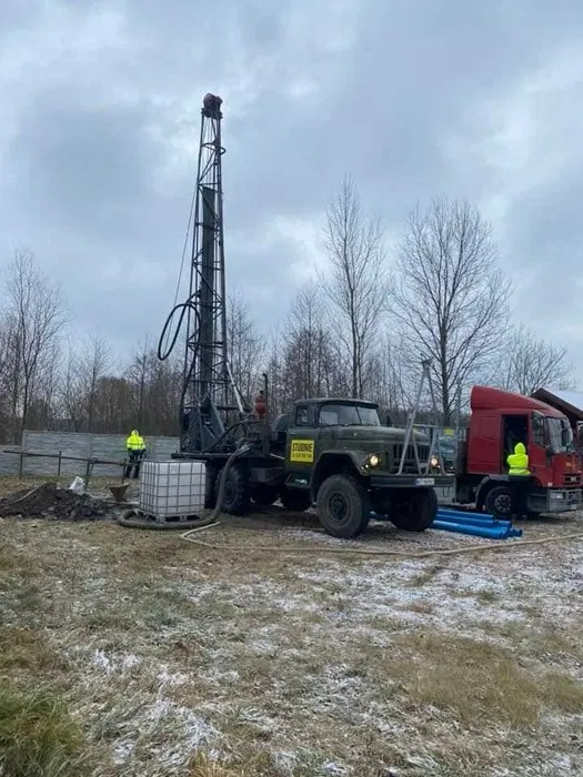 Mobilna wiertnica na ciężarówce podczas pracy na zewnątrz, widoczny pracownik w odblaskowej kamizelce i biały zbiornik IBC, w tle czerwona ciężarówka i drzewa bez liści.