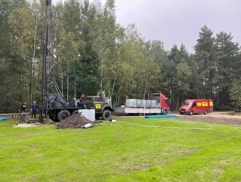 Wiertnica na podwoziu ciężarówki, dwóch pracowników przy pracy, obok ciężarówka z pojemnikami na wodę i czerwony bus z logo firmy na tle drzew i zielonej trawy.