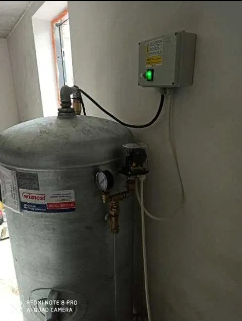 Szary zbiornik ciśnieniowy z manometrem i podłączeniami hydraulicznymi, zasilany z puszki elektrycznej z zieloną diodą, widoczny w jasnym pomieszczeniu.