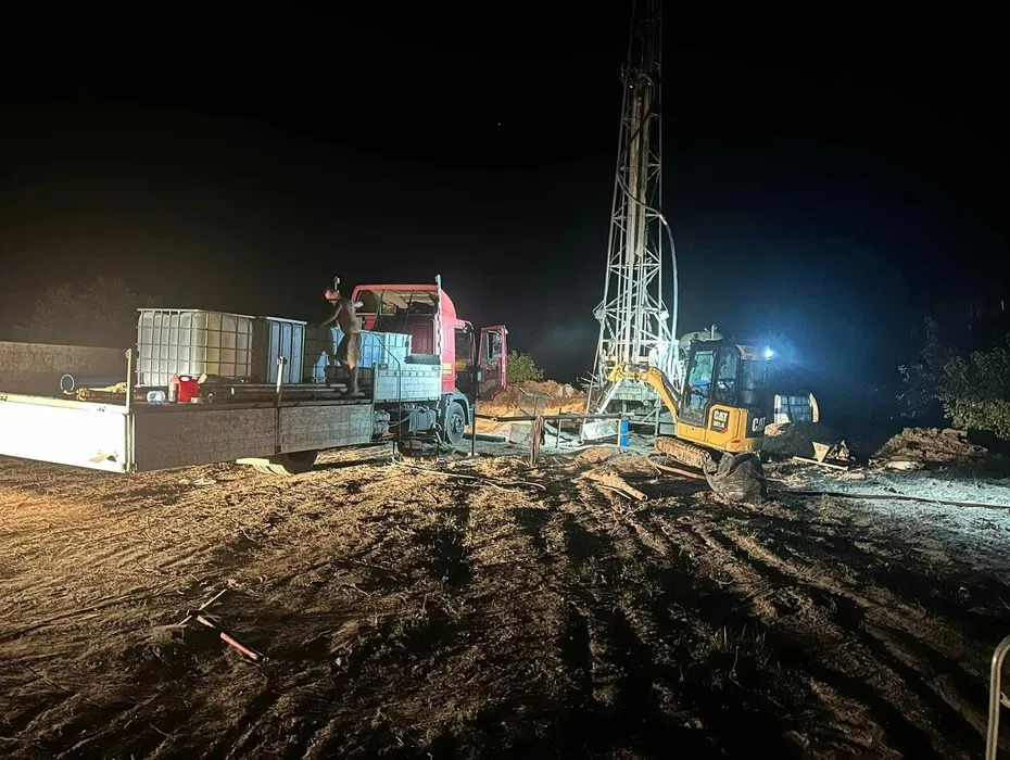 Prace wiertnicze nocą: widoczna wieża wiertnicza oświetlona reflektorami, obok koparka CAT i ciężarówka z pracownikiem na pace, teren błotnisty.