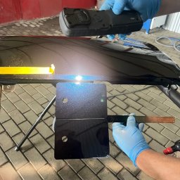 Lakiernia samochodowa Na Lwowskiej - Czarny element karoserii samochodu z przyklejoną żółtą taśmą, obok próbki lakieru z efektem brokatu, sprawdzane urządzeniem do pomiaru koloru przez osobę w niebieskich rękawiczkach.