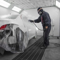 Lakiernia samochodowa Na Lwowskiej - Lakiernik w kombinezonie ochronnym, malujący biały samochód w komorze lakierniczej, z widoczną folią ochronną na pojeździe i kratką podłogową.