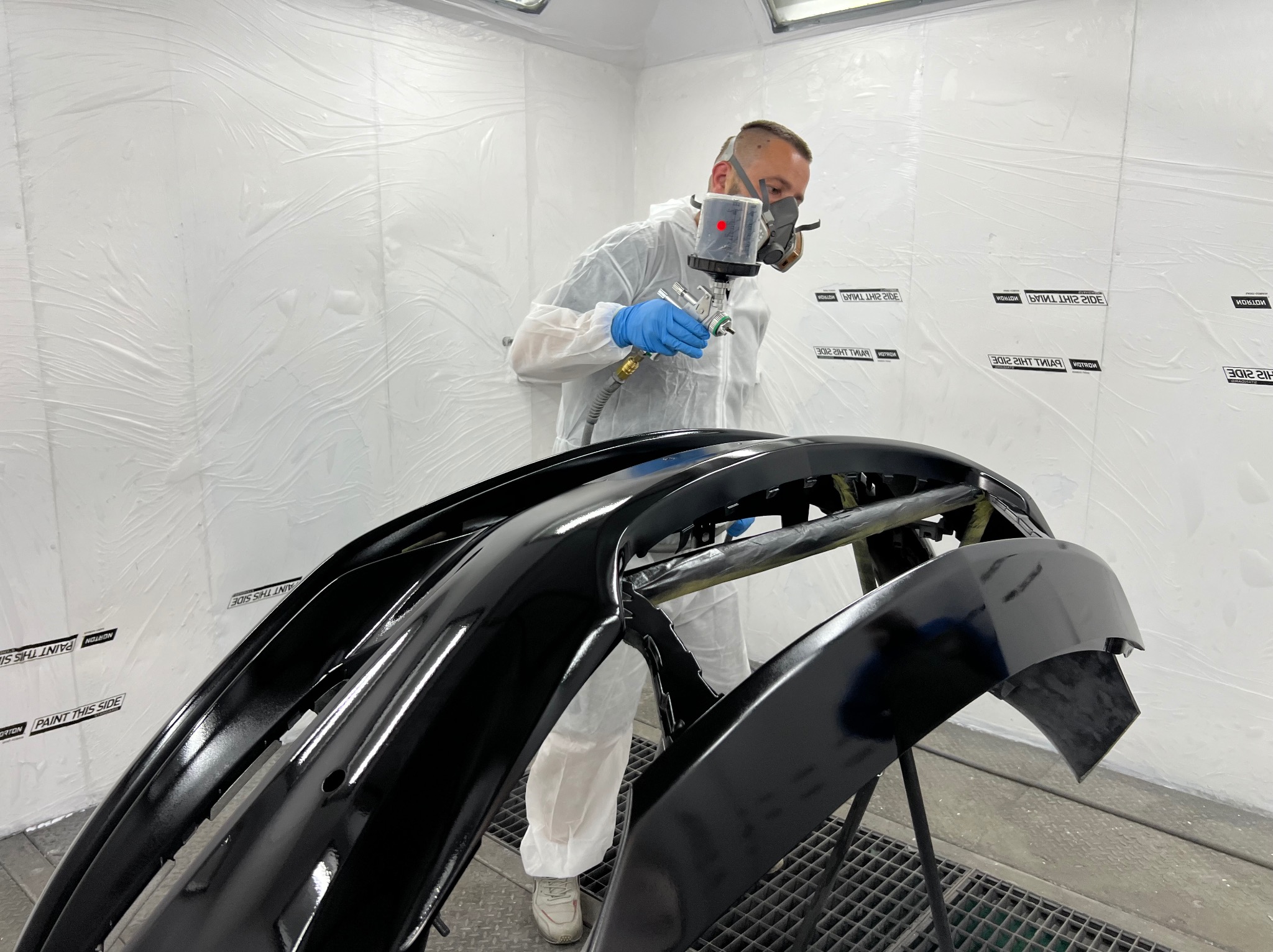 Lakiernik w kombinezonie ochronnym i masce maluje czarny zderzak samochodowy w kabinie lakierniczej, widoczne detale mocowania zderzaka.