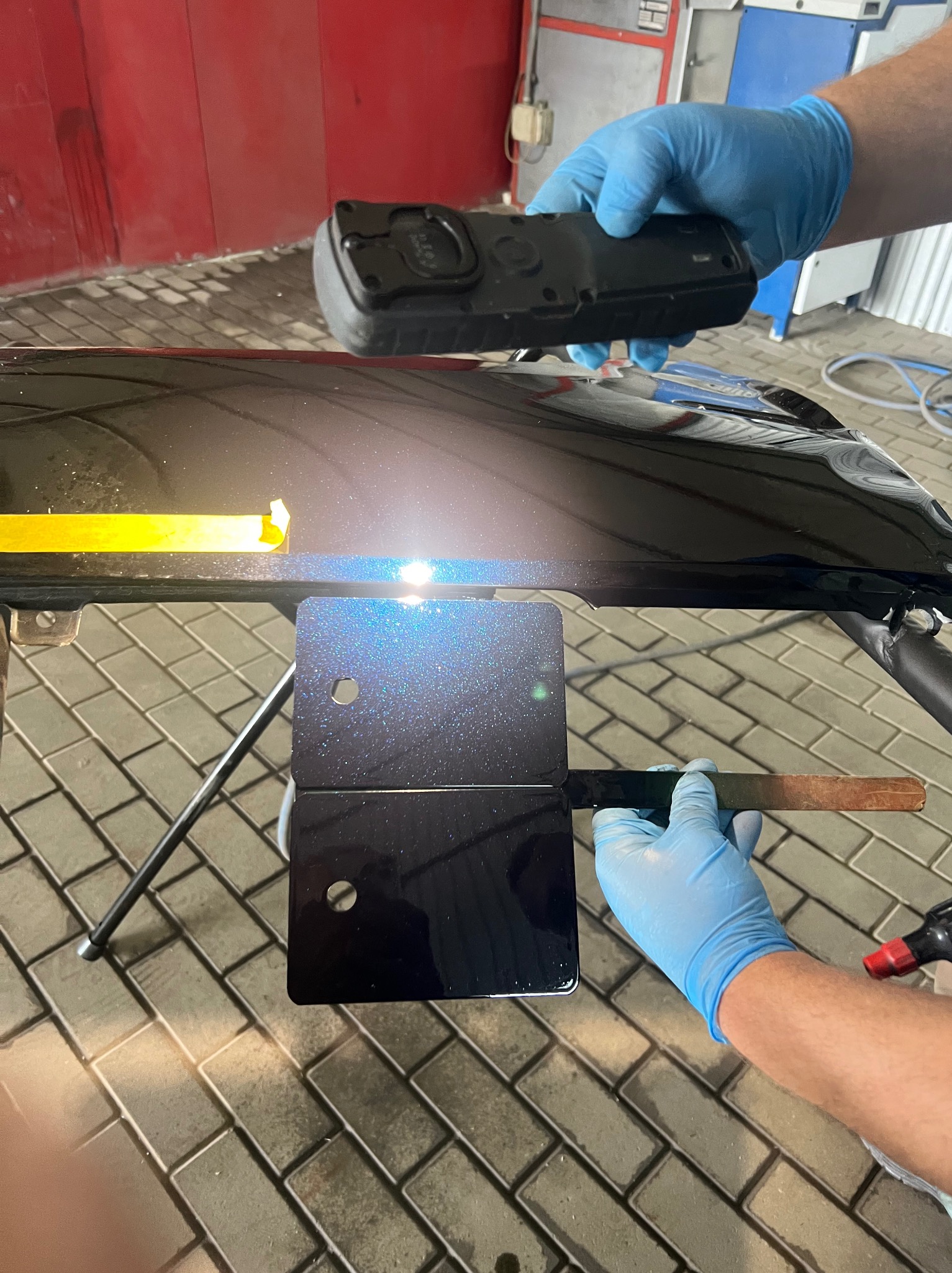 Czarny element karoserii samochodu z przyklejoną żółtą taśmą, obok próbki lakieru z efektem brokatu, sprawdzane urządzeniem do pomiaru koloru przez osobę w niebieskich rękawiczkach.