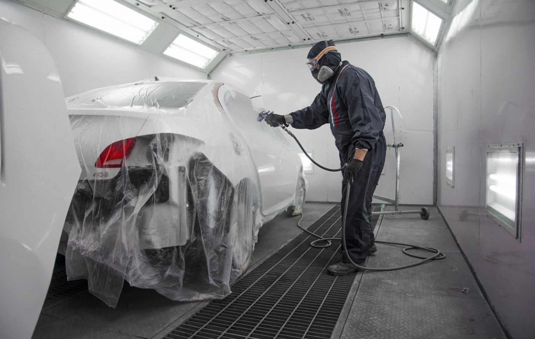 Lakiernik w kombinezonie ochronnym, malujący biały samochód w komorze lakierniczej, z widoczną folią ochronną na pojeździe i kratką podłogową.