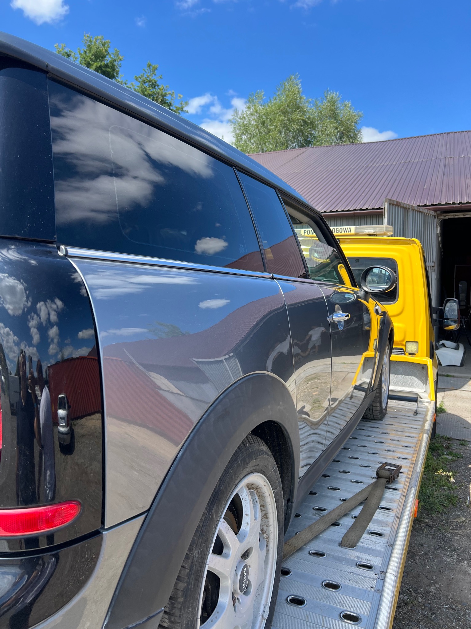 Czarno-szary Mini Clubman przewożony na żółtej lawecie, widoczne odbicie chmur na karoserii i aluminiowe felgi.