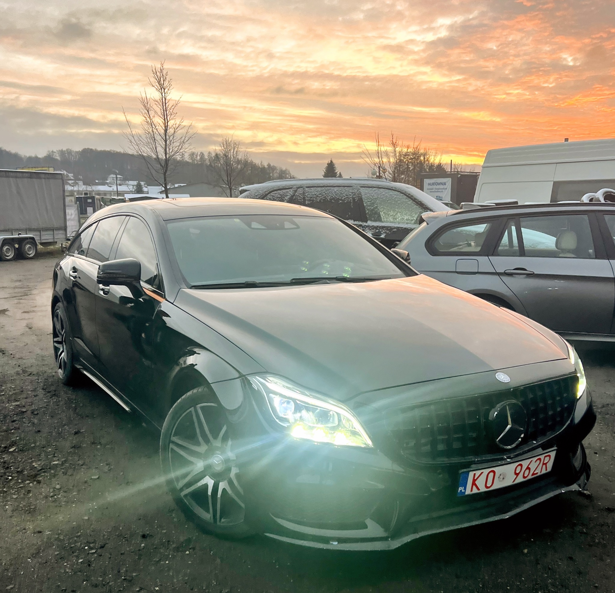 Czarny Mercedes-Benz CLS Shooting Brake z tuningowanym grillem i włączonymi światłami, zaparkowany na placu o zmierzchu.