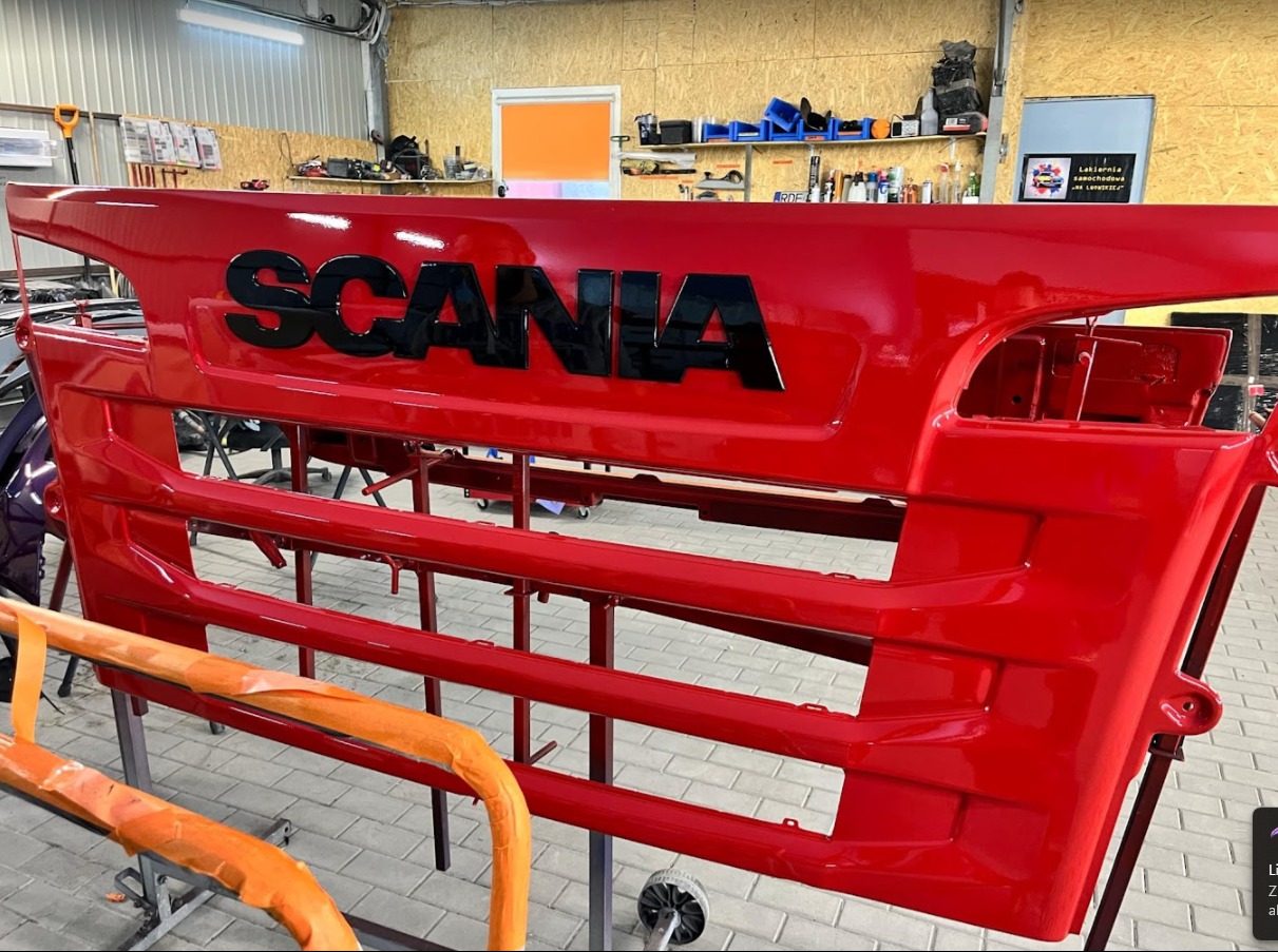 Czerwony grill ciężarówki Scania po lakierowaniu, zawieszony na stojaku w warsztacie, z widocznym napisem SCANIA w czarnym kolorze.