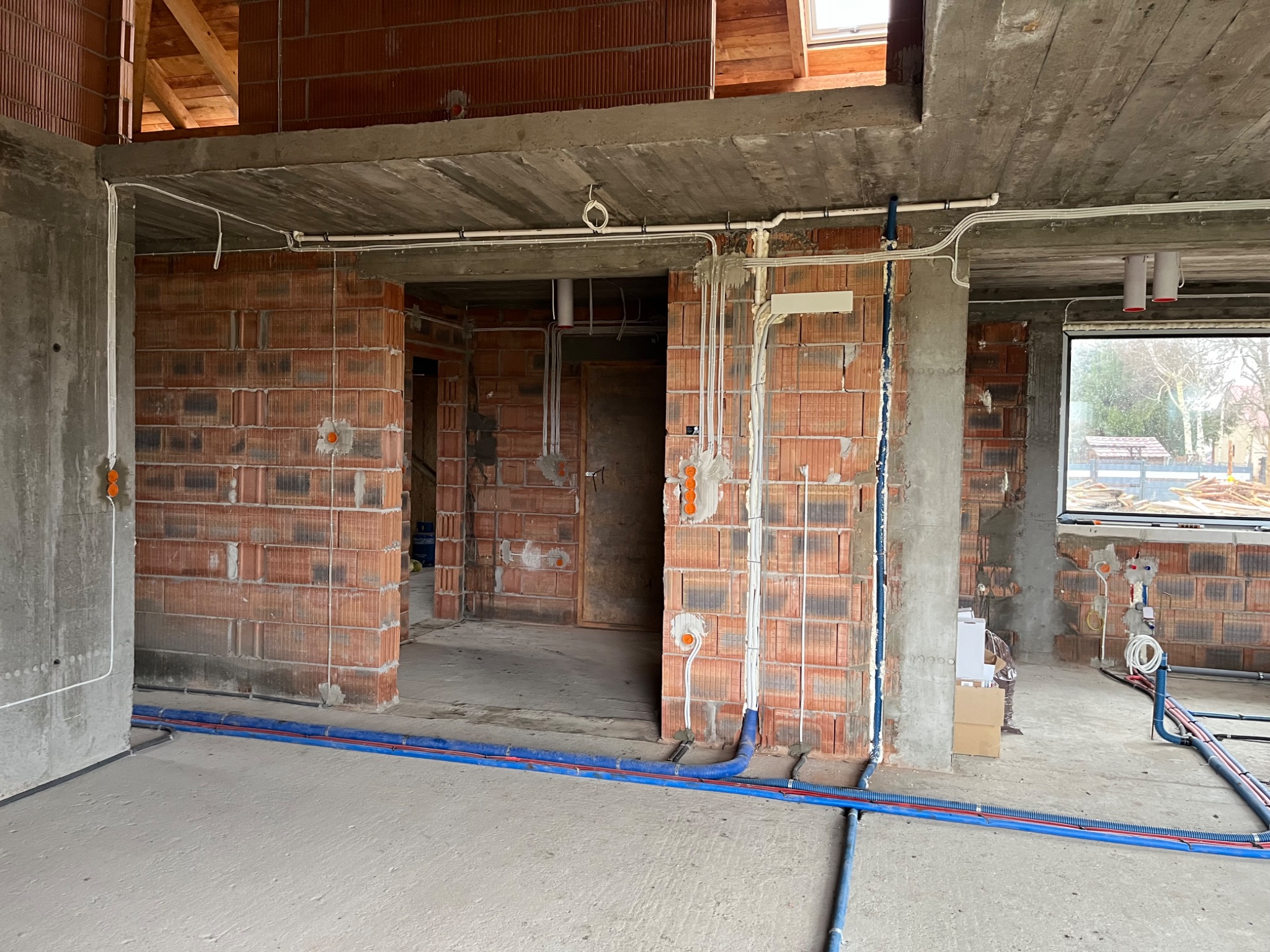Wnętrze w stanie surowym z widocznymi instalacjami elektrycznymi i hydraulicznymi, ceglane ściany, betonowy strop z widocznymi przewodami i rurami, okno z widokiem na zewnątrz.