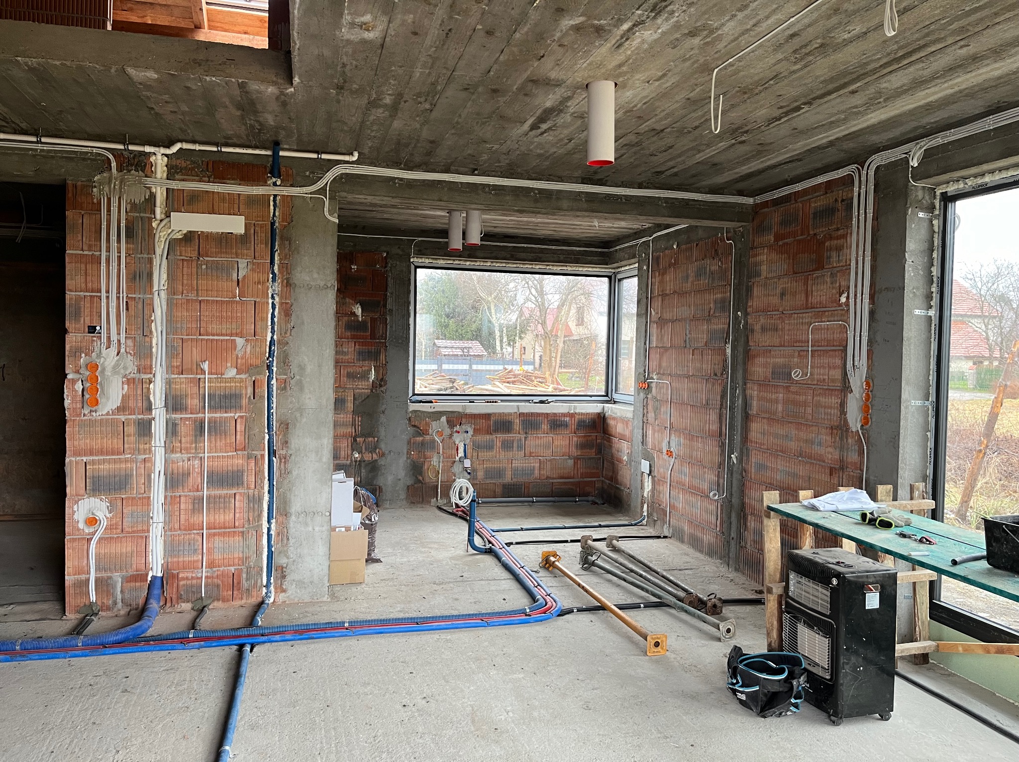 Wnętrze surowego domu w trakcie prac instalacyjnych: widoczne przewody elektryczne i rury rozprowadzone po ścianach z cegły, betonowy sufit z wystającymi oprawami oświetleniowymi, na podłodze...