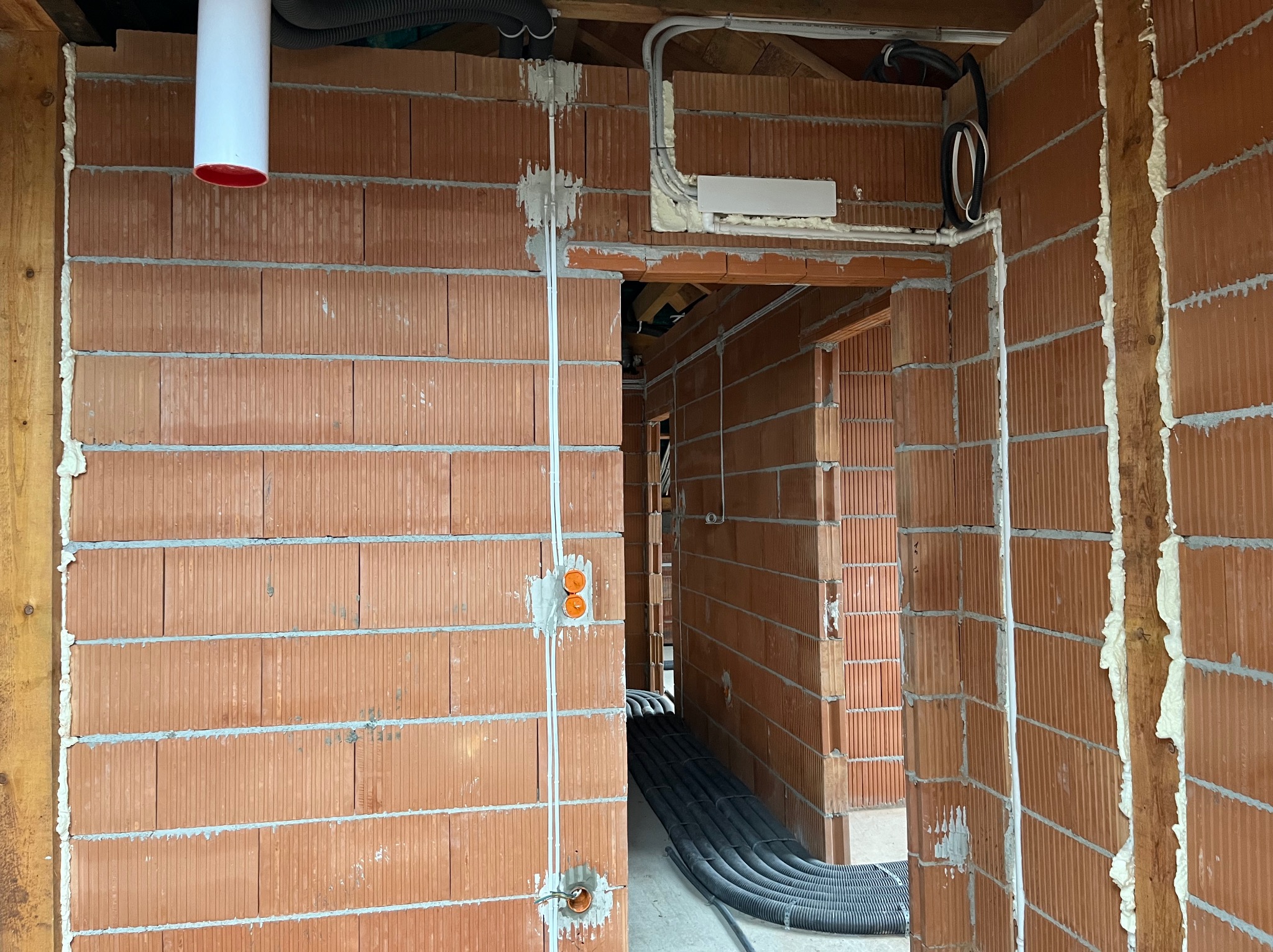 Wnętrze budynku w trakcie budowy z widocznymi przewodami elektrycznymi w ścianach z czerwonej cegły oraz rurami ochronnymi na podłodze.