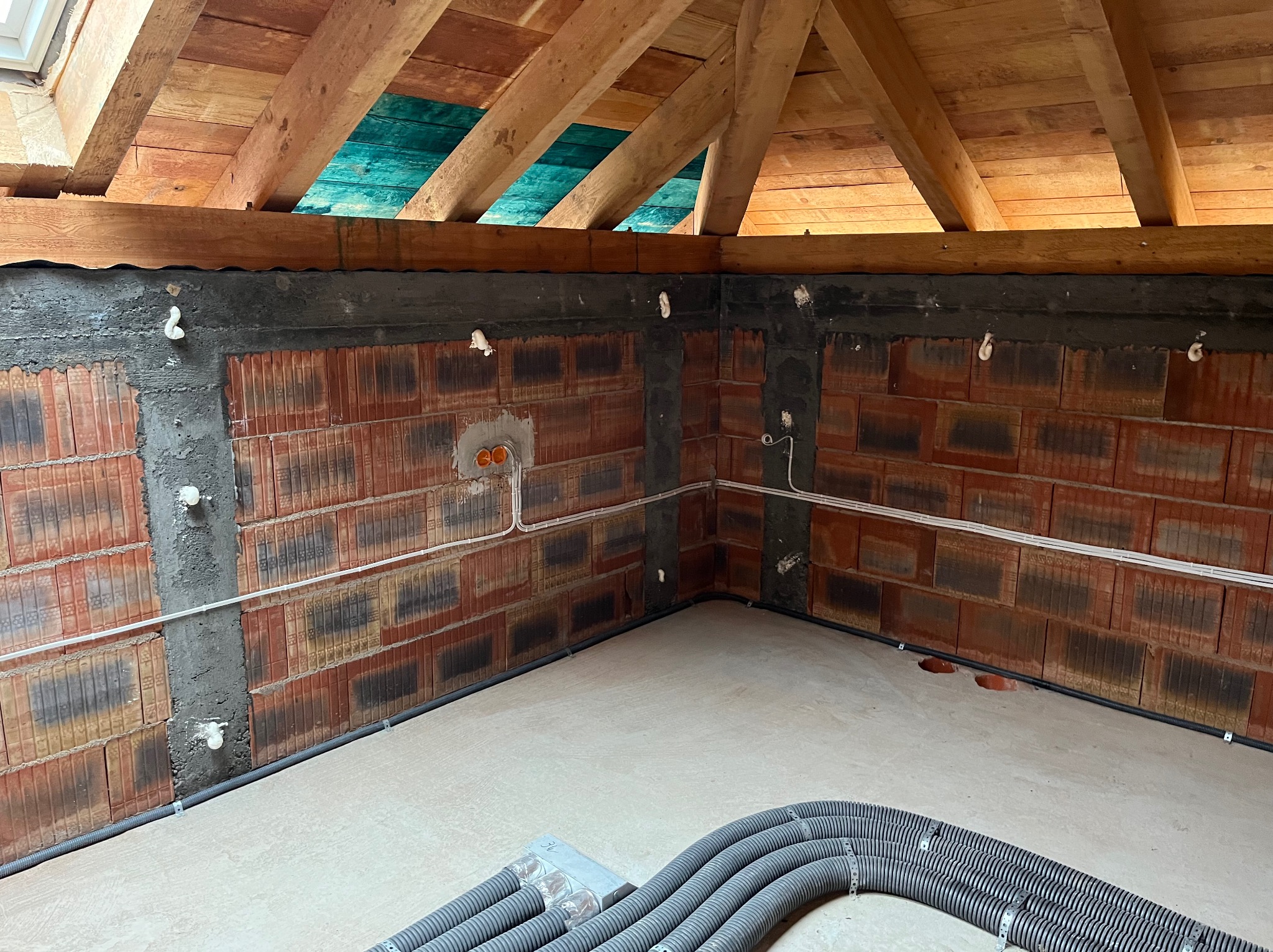 Wnętrze poddasza w trakcie prac elektrycznych: widoczne kable w peszlach, puszki instalacyjne w ścianie z cegły dziurawki, drewniana konstrukcja dachu z oknem dachowym.
