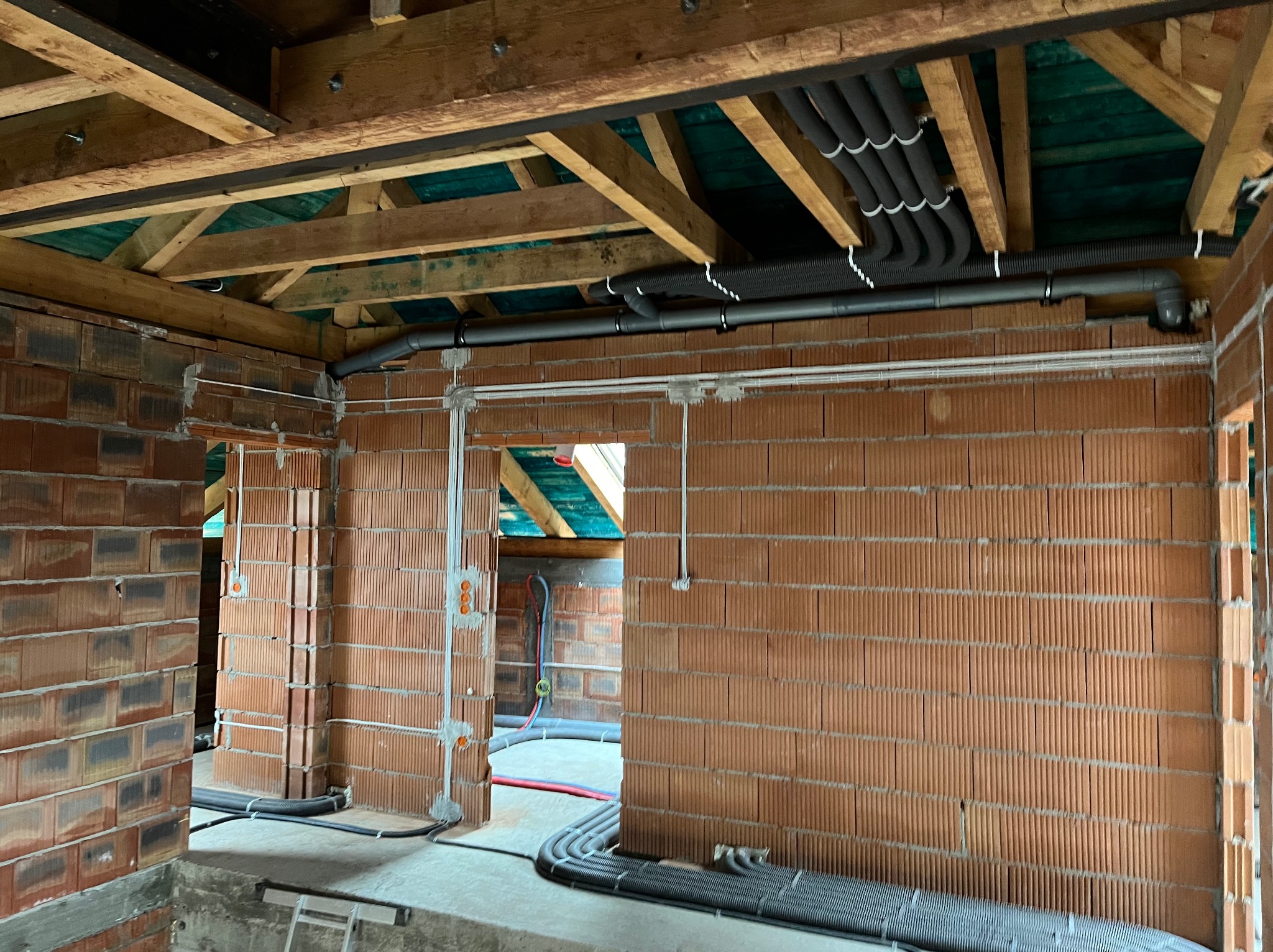 Surowy stan budynku w trakcie instalacji elektrycznej: widoczne kable w peszlach prowadzone po ścianach z cegły i pod sufitem z drewnianych belek.