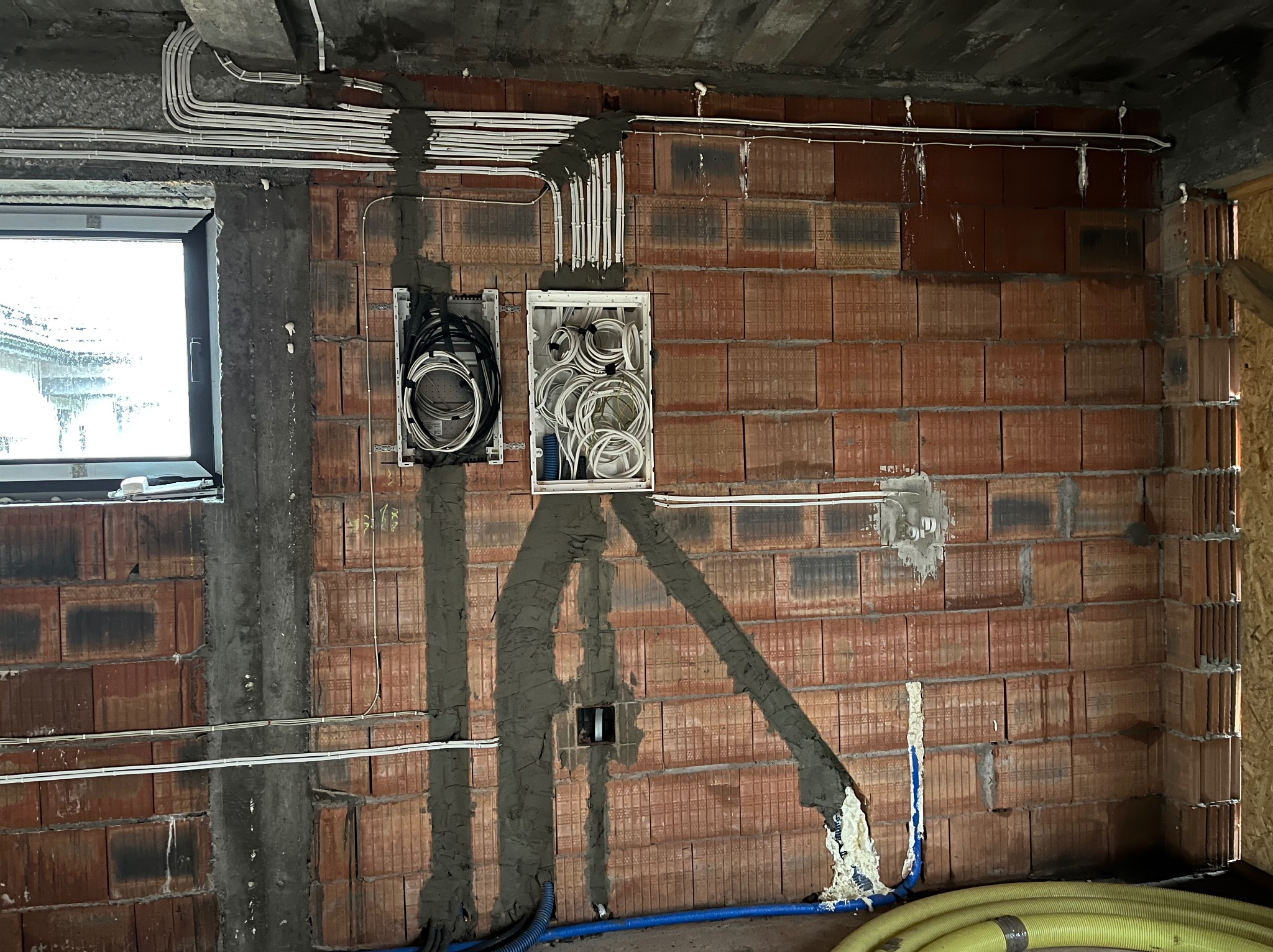 Wewnętrzna ściana z czerwonej cegły z zamontowanymi puszkami instalacyjnymi i białymi rurkami PCV prowadzącymi kable elektryczne; widoczne ślady świeżego tynku wokół puszek i rurek; okno w lewym...