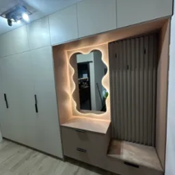 Beżowa szafa w przedpokoju z podświetlanym, nieregularnym lustrem, siedziskiem i szufladami, czarne uchwyty, panele ścienne z pionowych listew.