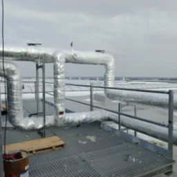 Izolowane rury instalacji na dachu budynku, widoczna krata pomostowa, paleta drewniana i wiadro z narzędziami, pochmurne niebo w tle.