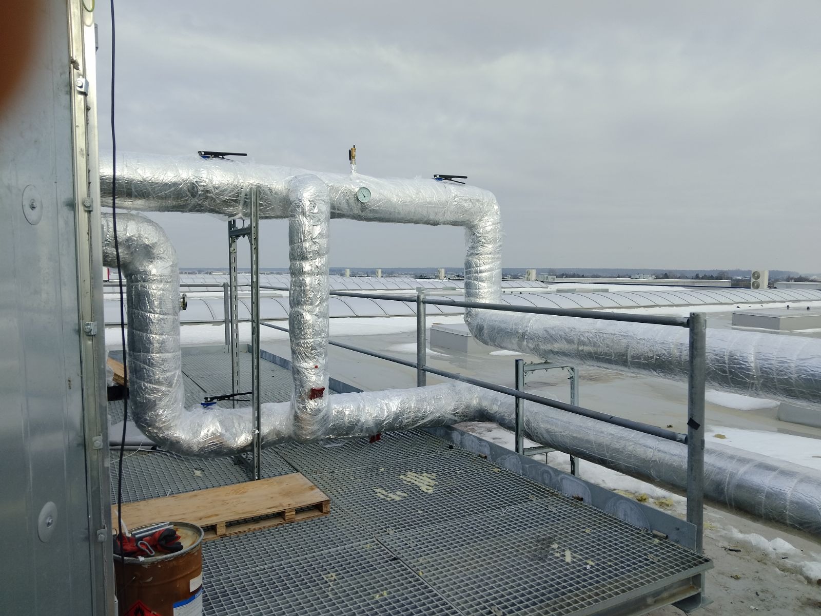 Izolowane rury instalacji na dachu budynku, widoczna krata pomostowa, paleta drewniana i wiadro z narzędziami, pochmurne niebo w tle.