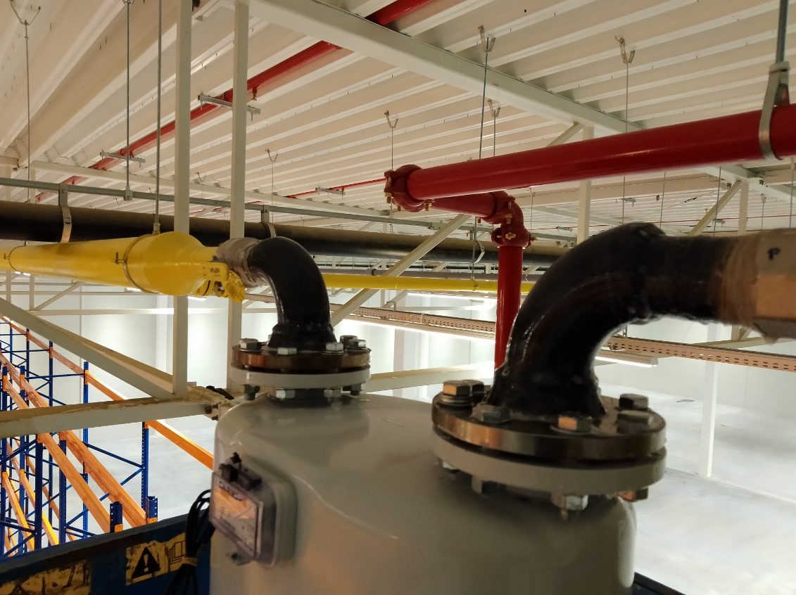 Widok na instalację przemysłową z rurami w kolorach żółtym, czerwonym i czarnym w hali w Łowinku, z widocznymi elementami mocującymi i zabezpieczeniami.