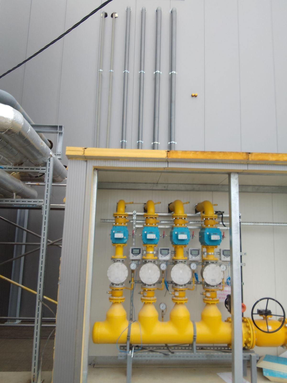 Żółta instalacja gazowa z czterema zaworami i manometrami wewnątrz metalowej konstrukcji na tle szarej ściany z rurami. Widoczny fragment koła zaworu.