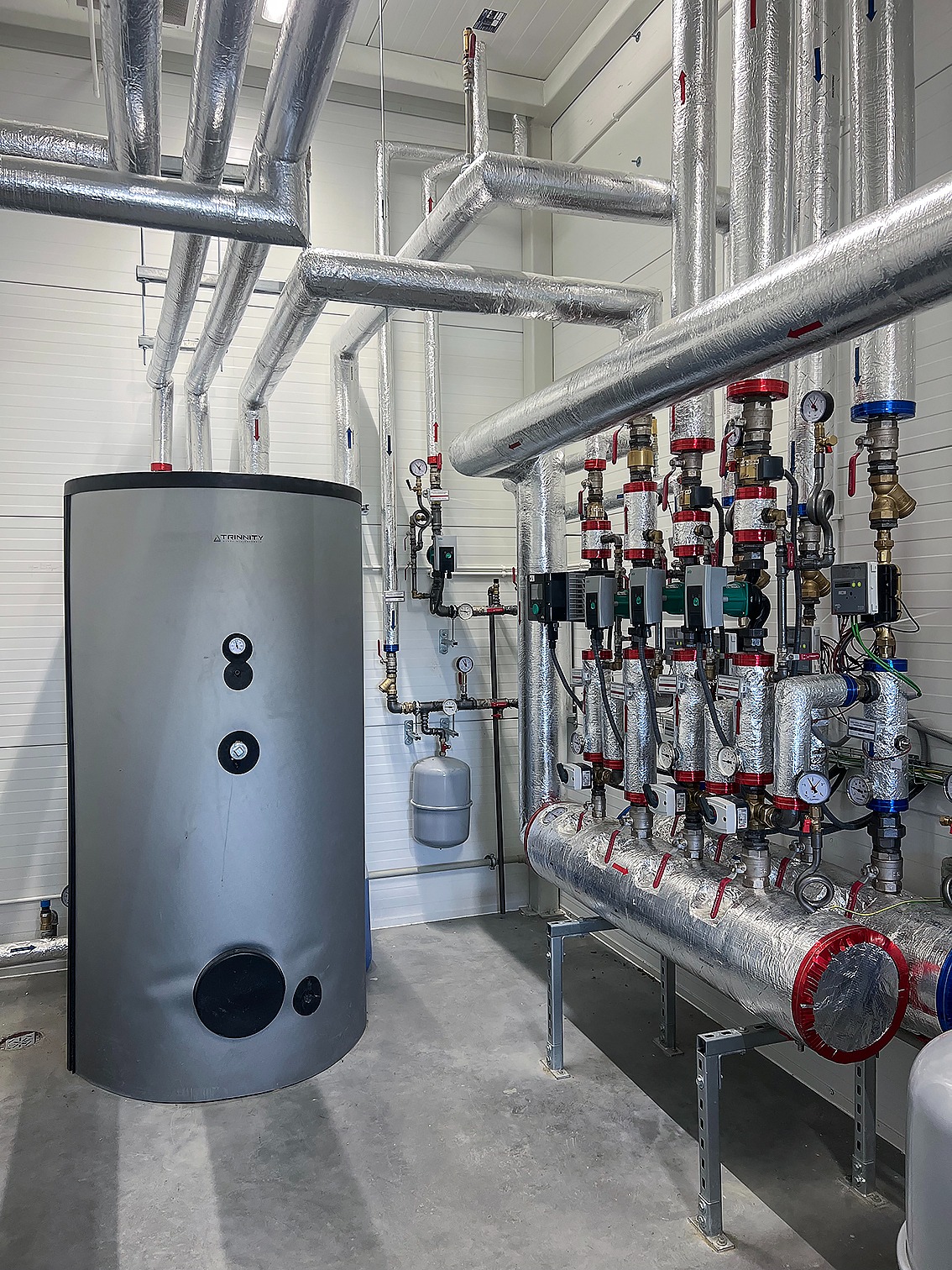 Szary piec CO marki Atrinnity z instalacją grzewczą w srebrnej izolacji, widoczne liczne zawory i manometry. Nowoczesna kotłownia w jasnym pomieszczeniu.