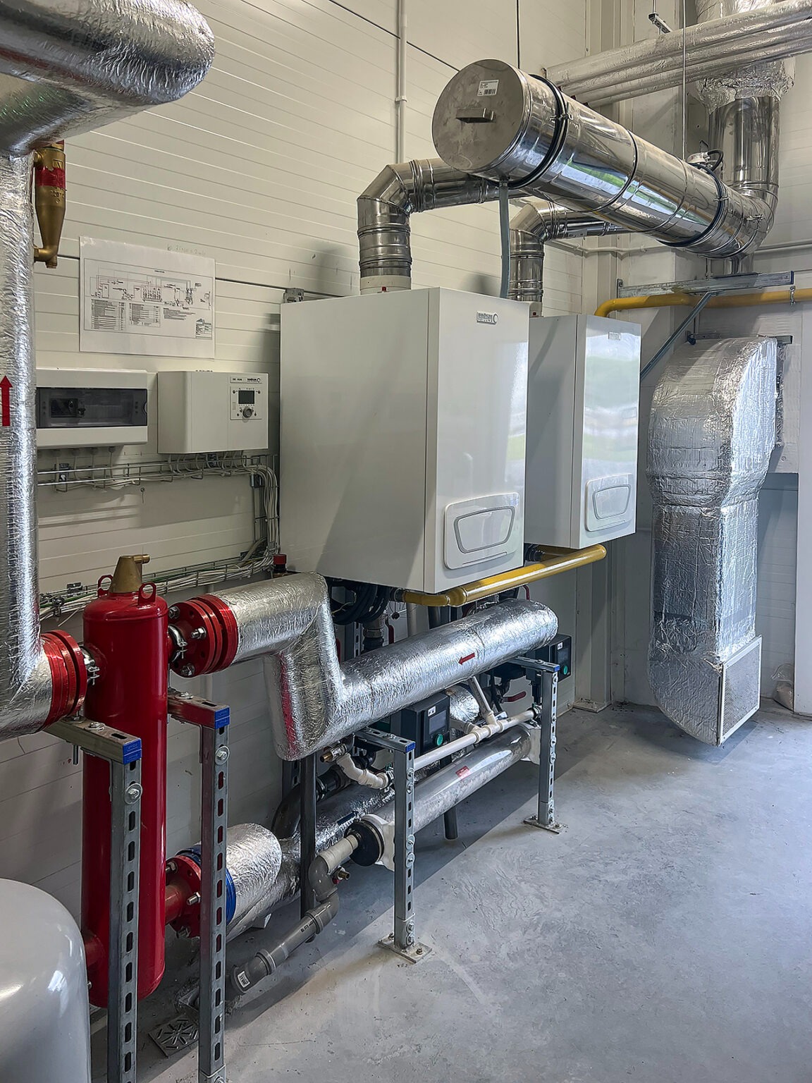 Dwa białe, wiszące piece gazowe z podłączeniami, rurami w srebrnej izolacji i czerwonym zaworem. Na ścianie schemat instalacji. Nowoczesna kotłownia.