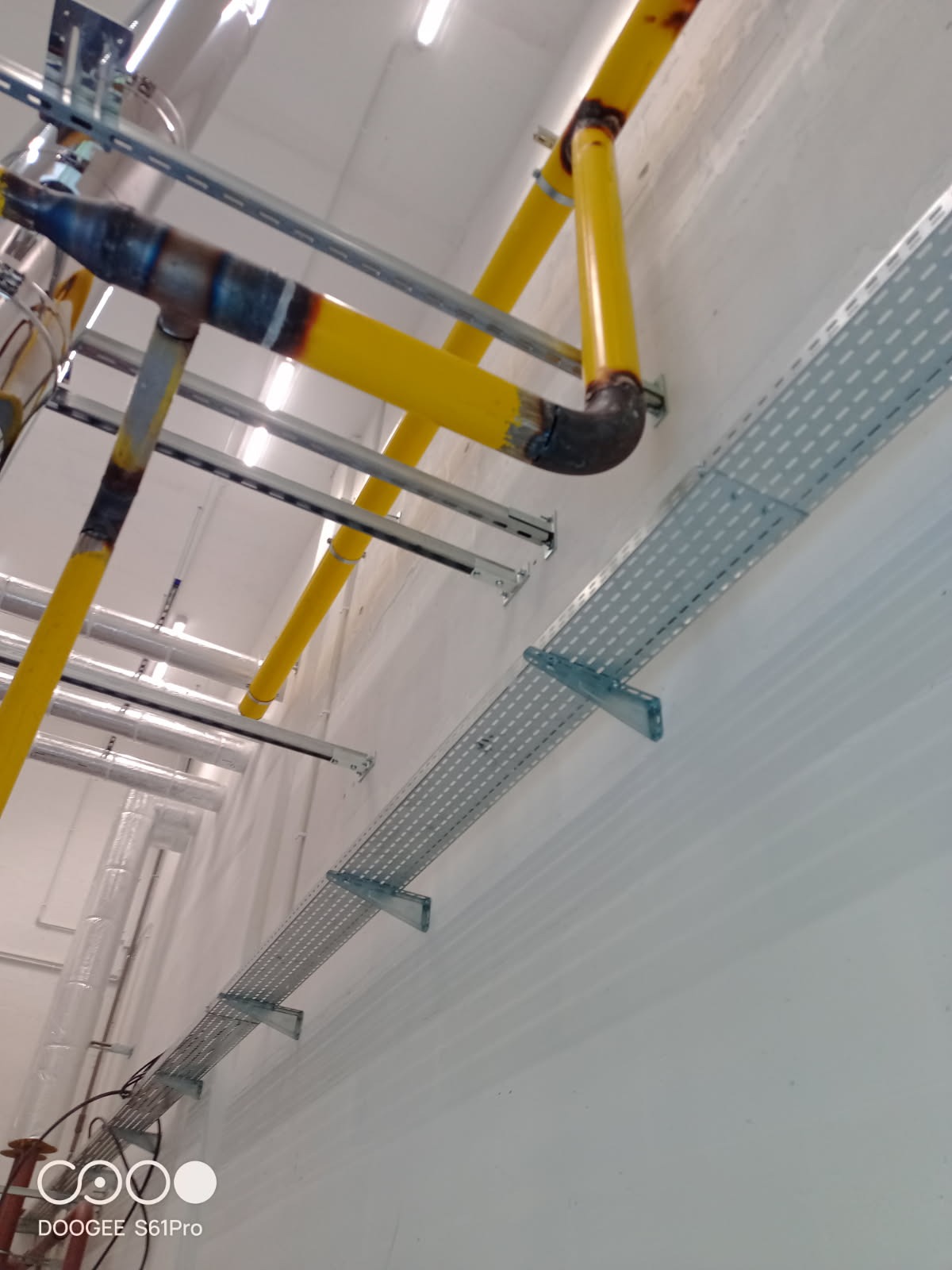 Żółte rury instalacyjne z widocznymi spawami, zamocowane do ściany na metalowych wspornikach. Obok koryto kablowe. Ujęcie z dołu, widoczny fragment sufitu.