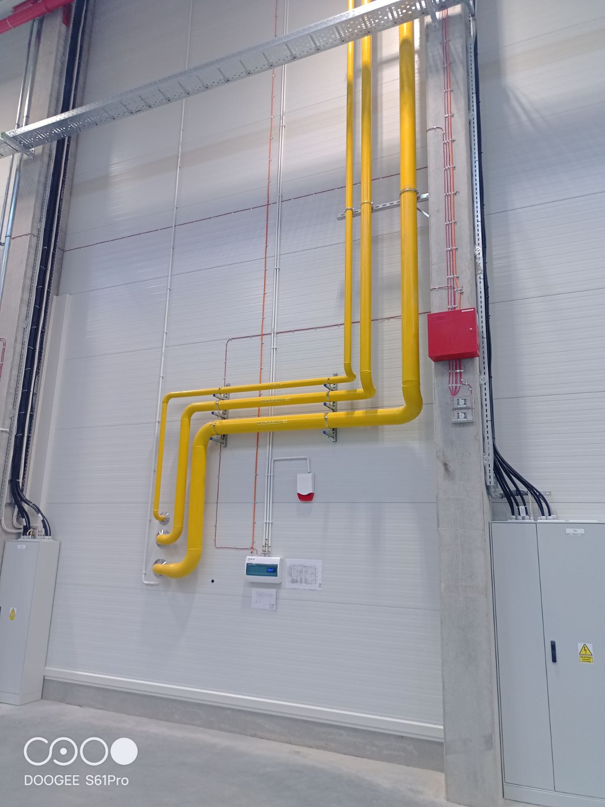 Żółte rury gazowe zamontowane w hali przemysłowej, widoczne elementy instalacji elektrycznej i skrzynki. Białe ściany z paneli, szara podłoga.