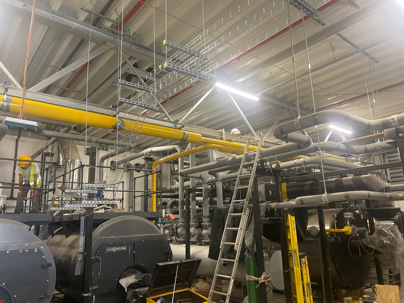 Rozbudowana instalacja przemysłowa z rurami, kotłami Viessmann i drabina. Pracownik w kasku i kamizelce odblaskowej. Widok na sufit z instalacjami.
