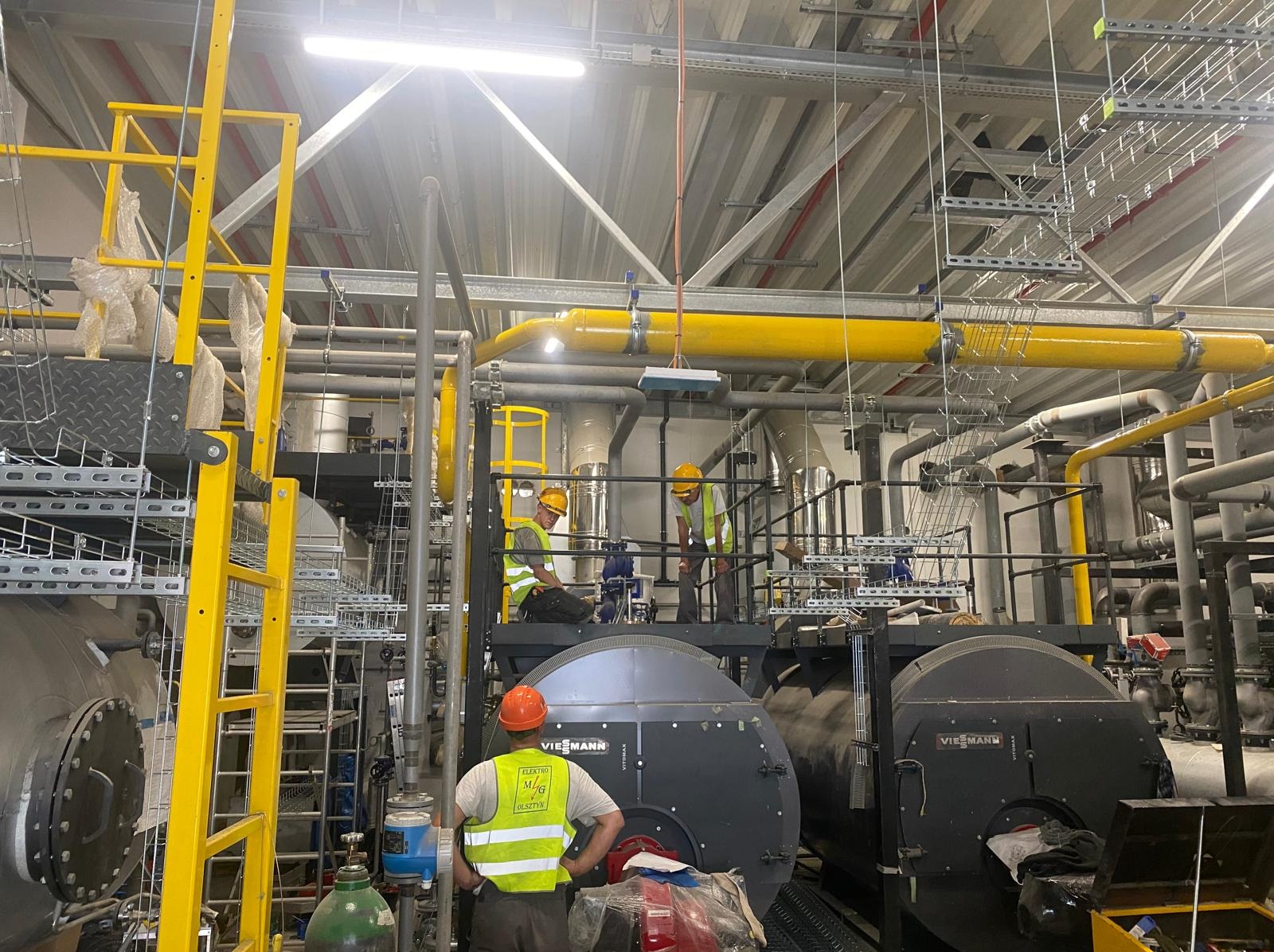 Montaż kotłów Viessmann w kotłowni przemysłowej. Żółte rury, drabiny i pracownicy w kaskach. Jeden z pracowników stoi tyłem, widoczny napis na kamizelce.