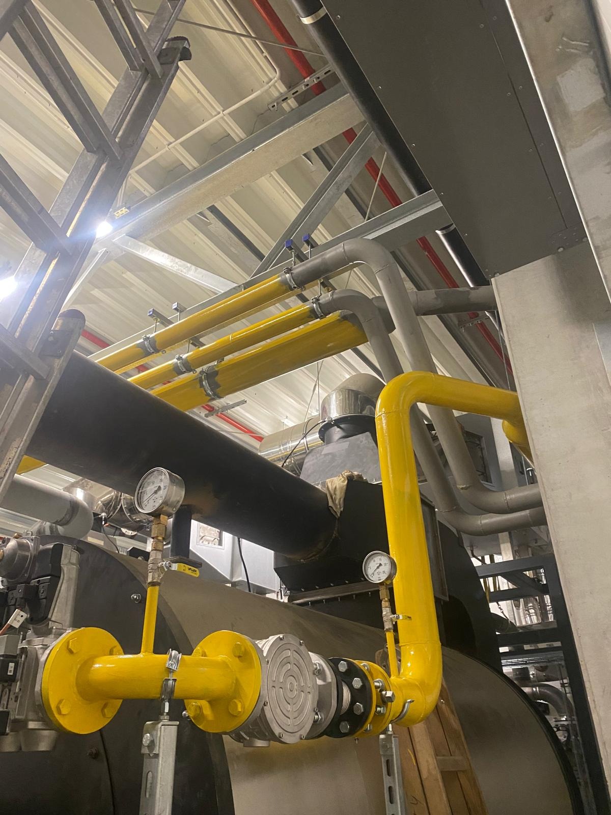 Kompleksowa instalacja przemysłowa z rurami gazowymi w żółtej izolacji, manometry i elementy mocujące. Widok z dołu na konstrukcję pod stropem.