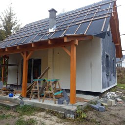 Fasad Servis - Budowa domu jednorodzinnego w Gdańsku: widok na elewację w trakcie ocieplania, z widocznym styropianem i przygotowaniem podłoża pod tynk, drewniana konstrukcja werandy z pomalowanymi słupami.