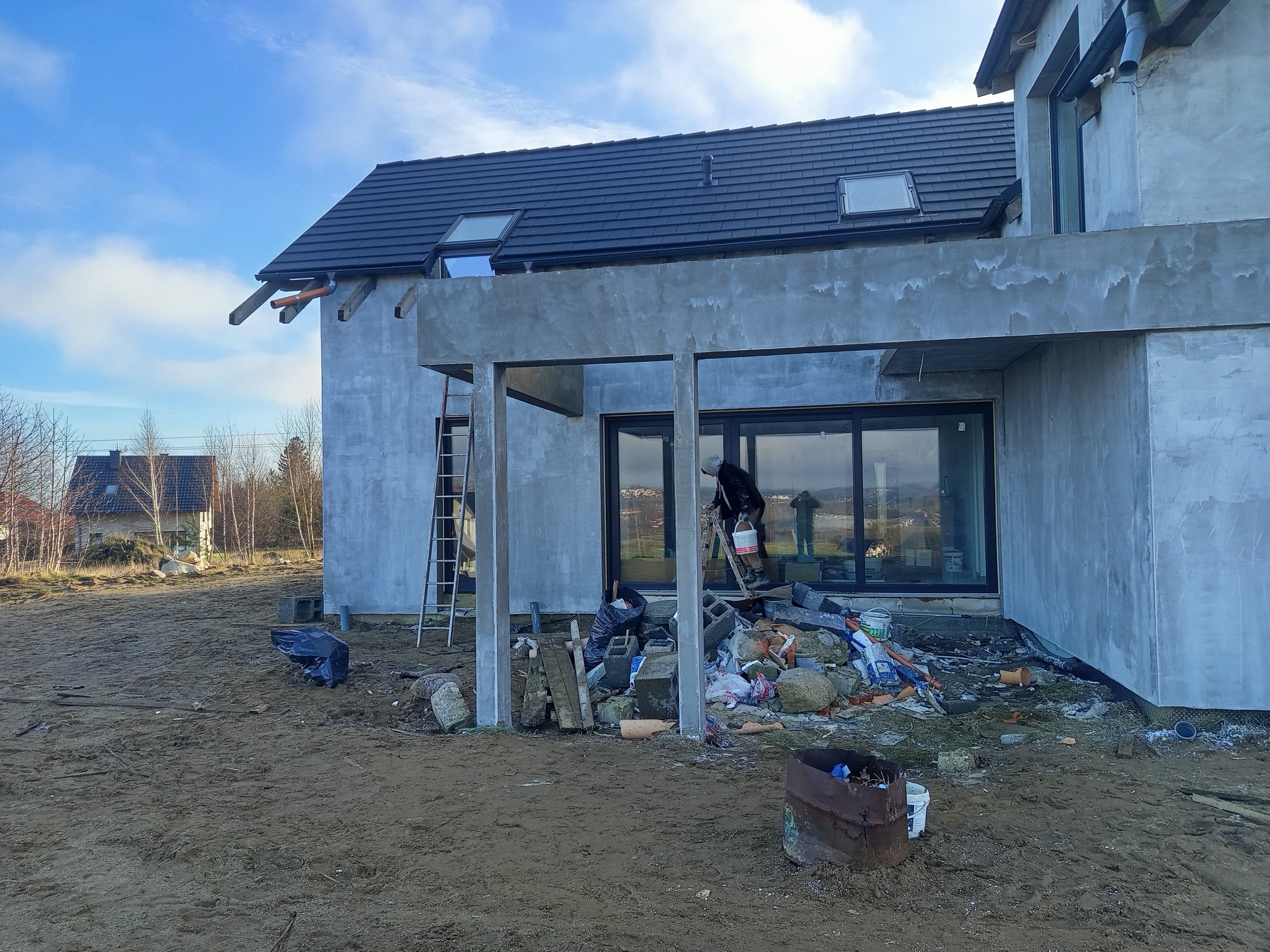 Surowa elewacja domu jednorodzinnego w trakcie prac wykończeniowych; widoczny pracownik z wiadrem na drabinie, materiały budowlane i bałagan na placu budowy