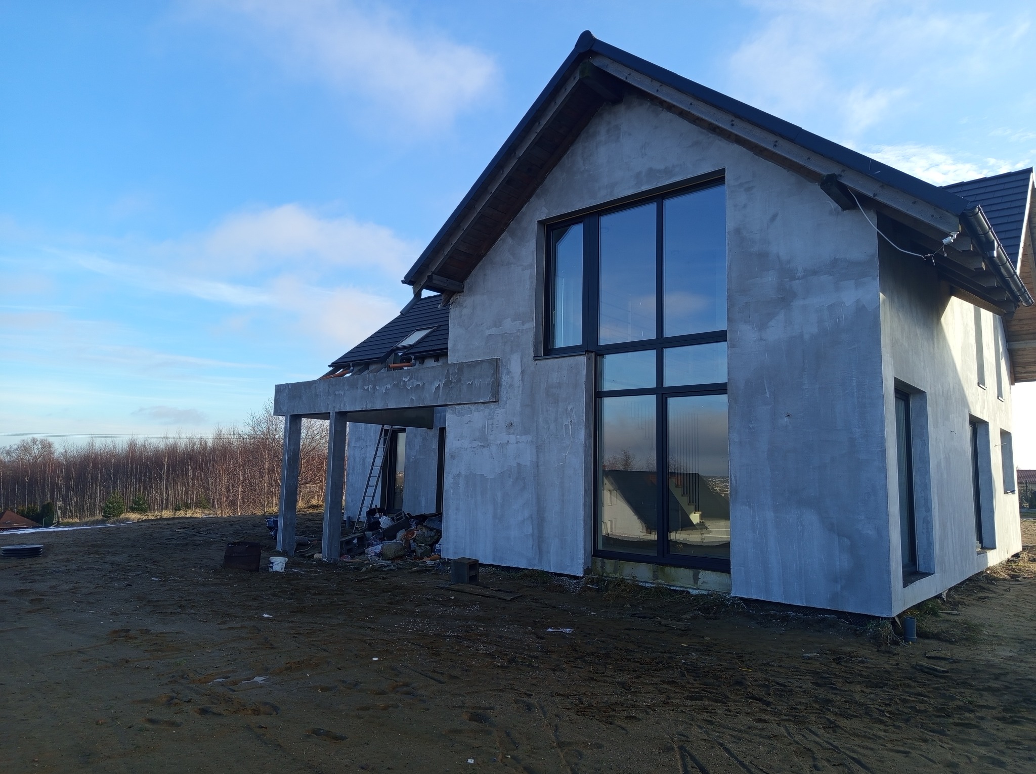 Dom jednorodzinny w trakcie prac elewacyjnych, widoczne szare tynki i duże okna z czarnymi ramami, teren wokół domu nierówny, częściowo pokryty ziemią.