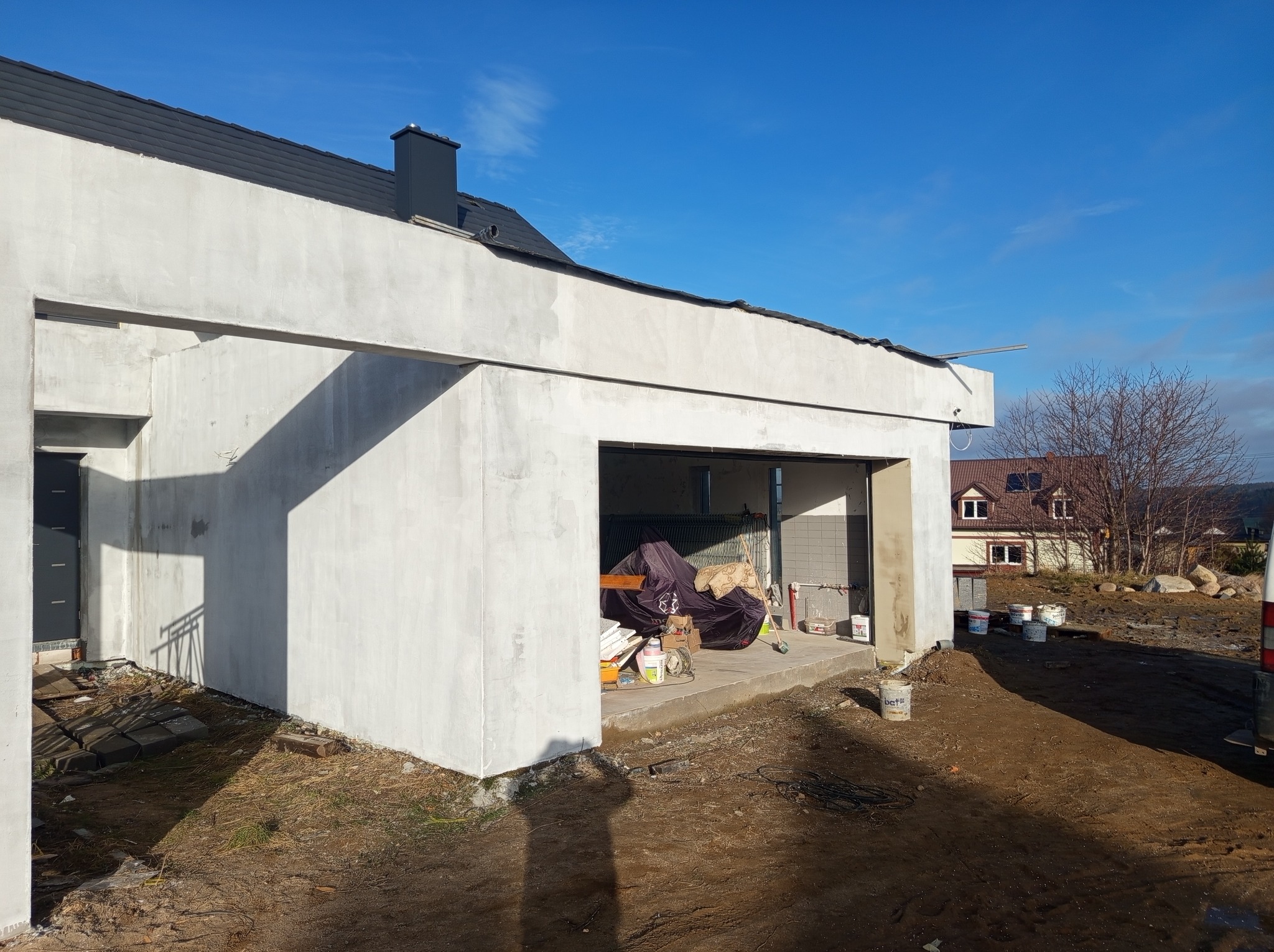 Budynek w trakcie wykonywania elewacji, z widocznymi śladami świeżego tynku, otwarty garaż z narzędziami i materiałami budowlanymi, ziemiste podłoże i błękitne niebo w tle.