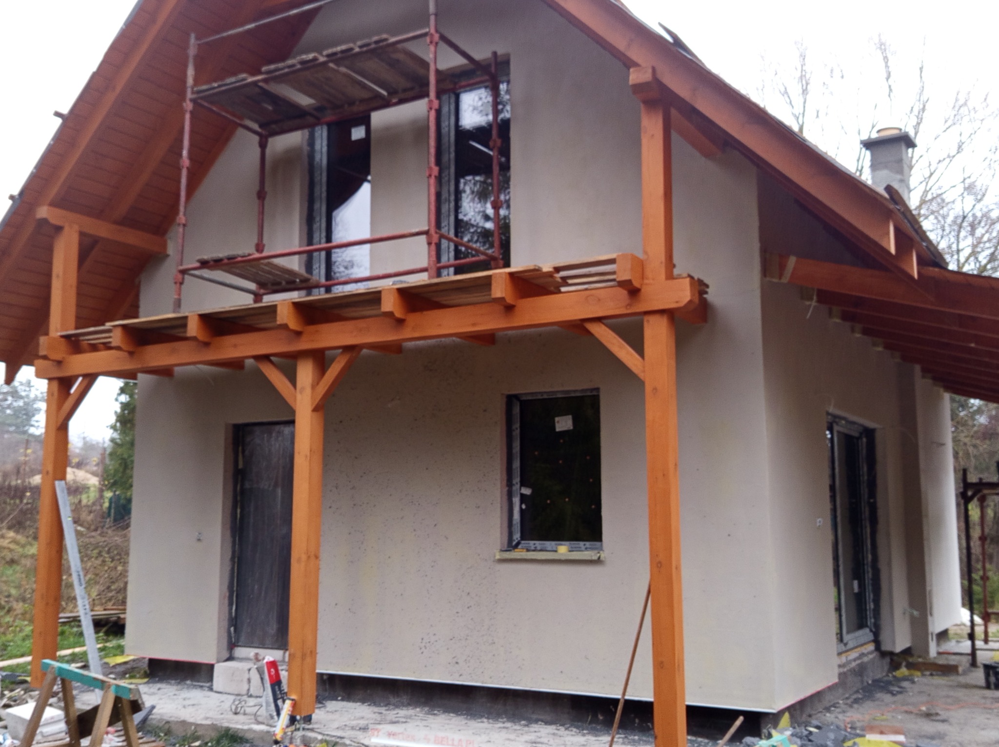 Dom jednorodzinny w trakcie prac elewacyjnych, widoczne rusztowania i świeżo nałożony tynk strukturalny w odcieniach beżu, drewniana konstrukcja zadaszenia.