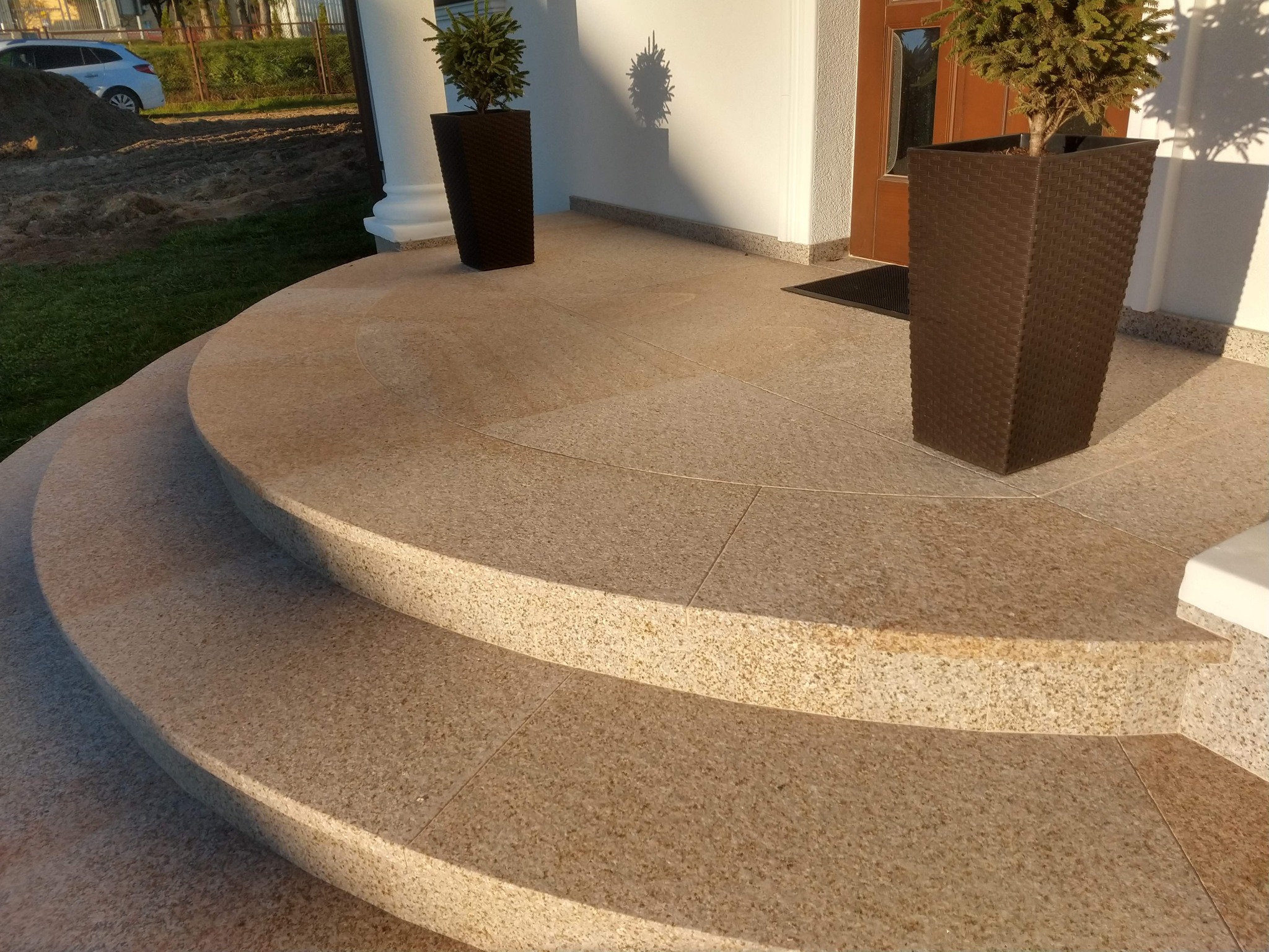 Półokrągłe wejściowe stopnie z jasnego, nakrapianego kamienia, z donicami z iglakami po bokach, prowadzące do drzwi wejściowych.