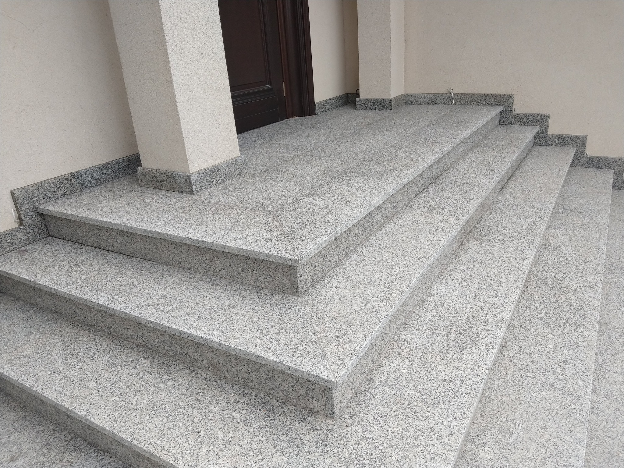 Zewnętrzne stopnie wejściowe wyłożone płytami z szarego, drobnoziarnistego kamienia z ciemnymi obrzeżami, prowadzące do brązowych drzwi.